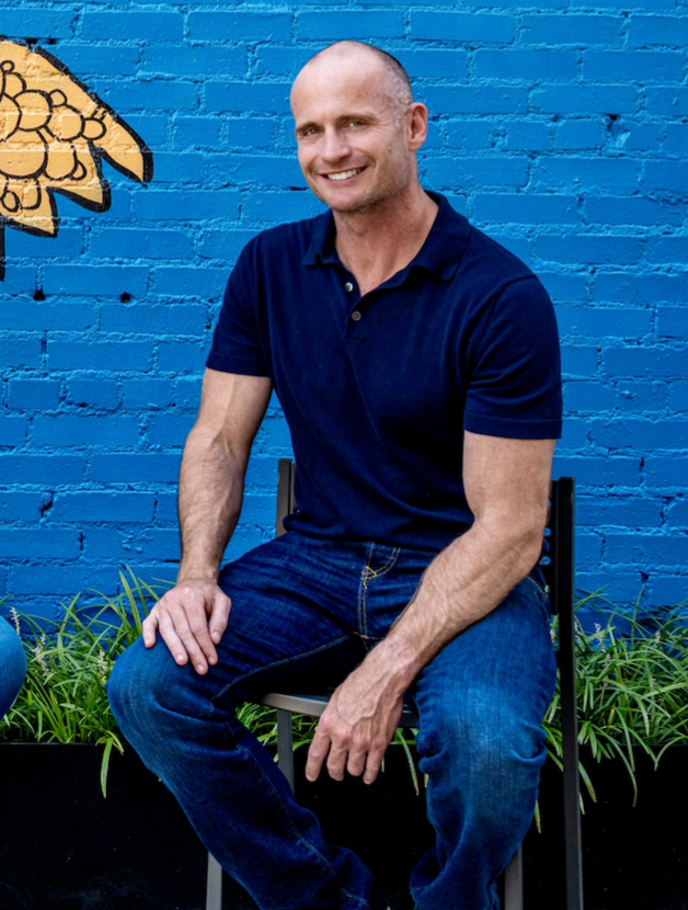 John Miller sitting on a stool in front of a blue wall with painted flowers, representing the founder of Agile Classrooms and Certified Scrum Trainer